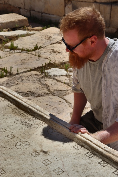A researcher examining an ancient carved stone surface featuring geometric patterns and game board markings at an archaeological site.