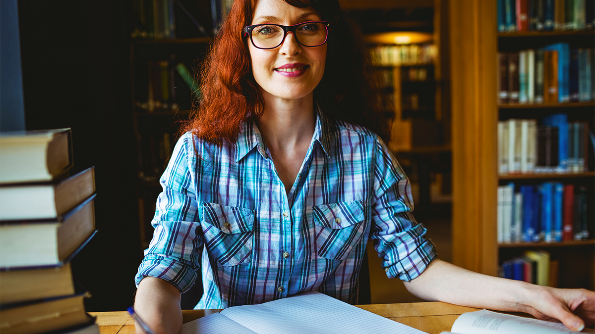 Female student sitting in library making notes at a desk with a pile of books next to her