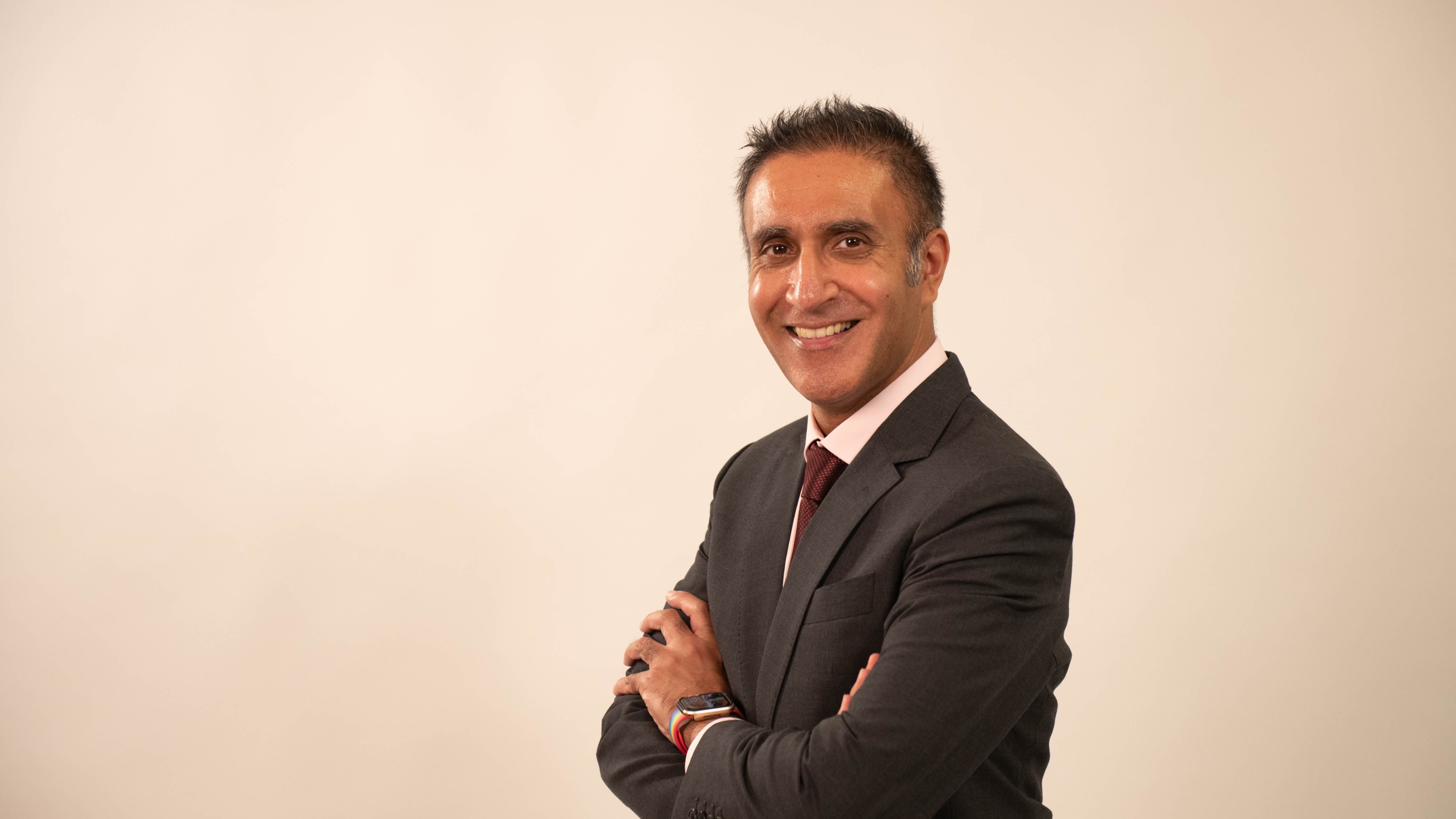 Jatinder, wearing a dark tailored suit, white shirt and tie stands at a slight angle against a light neutral background with arms folded and a smartwatch visible on one wrist.